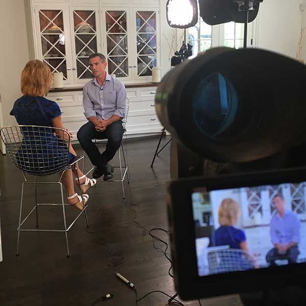 Sarah Wallace interviewing Fotis Dulos on camera in a white room, the scene visible in the camera viewfinder.