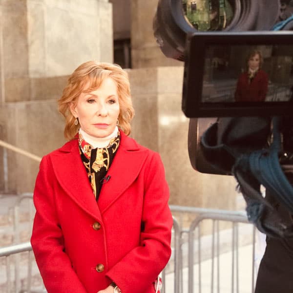 Sarah Wallace in a red coat on camera, her image reflected in the lens.